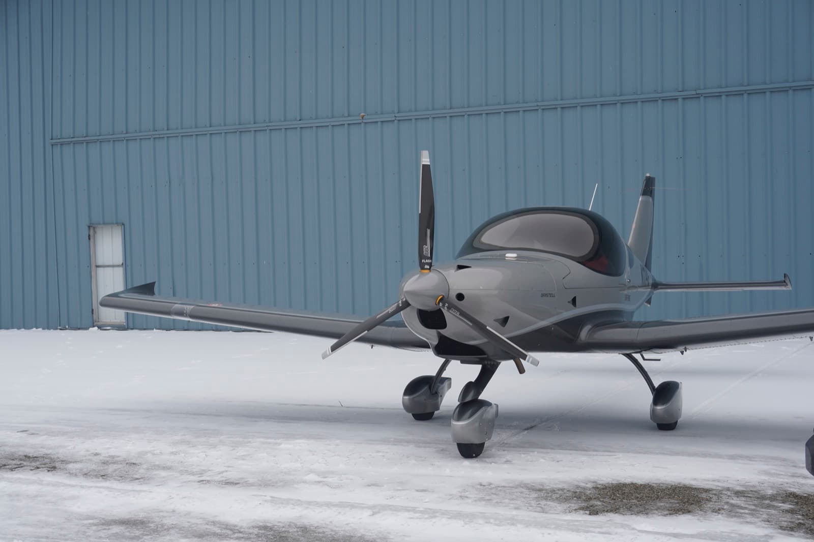 A silver Bristell parked alongside the hangar in winter light