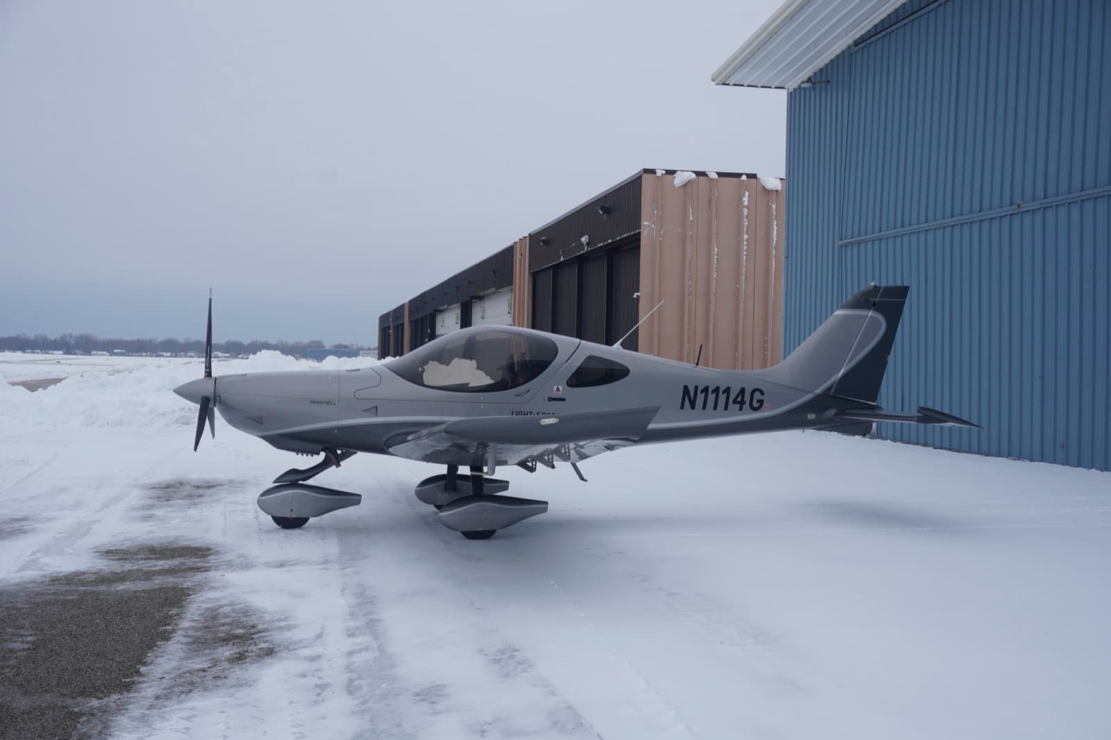 A silver Bristell beside the blue hangar after snowfall