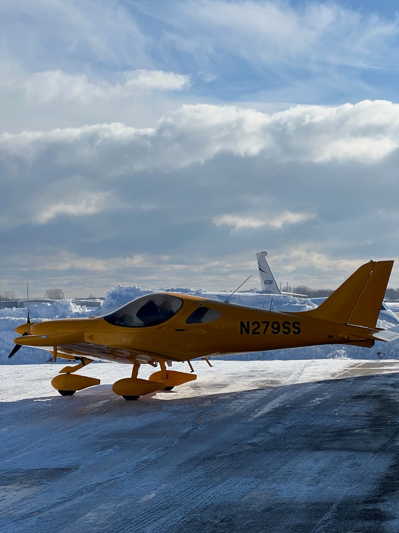 A yellow Bristell on a snow covered ramp under a broken sky