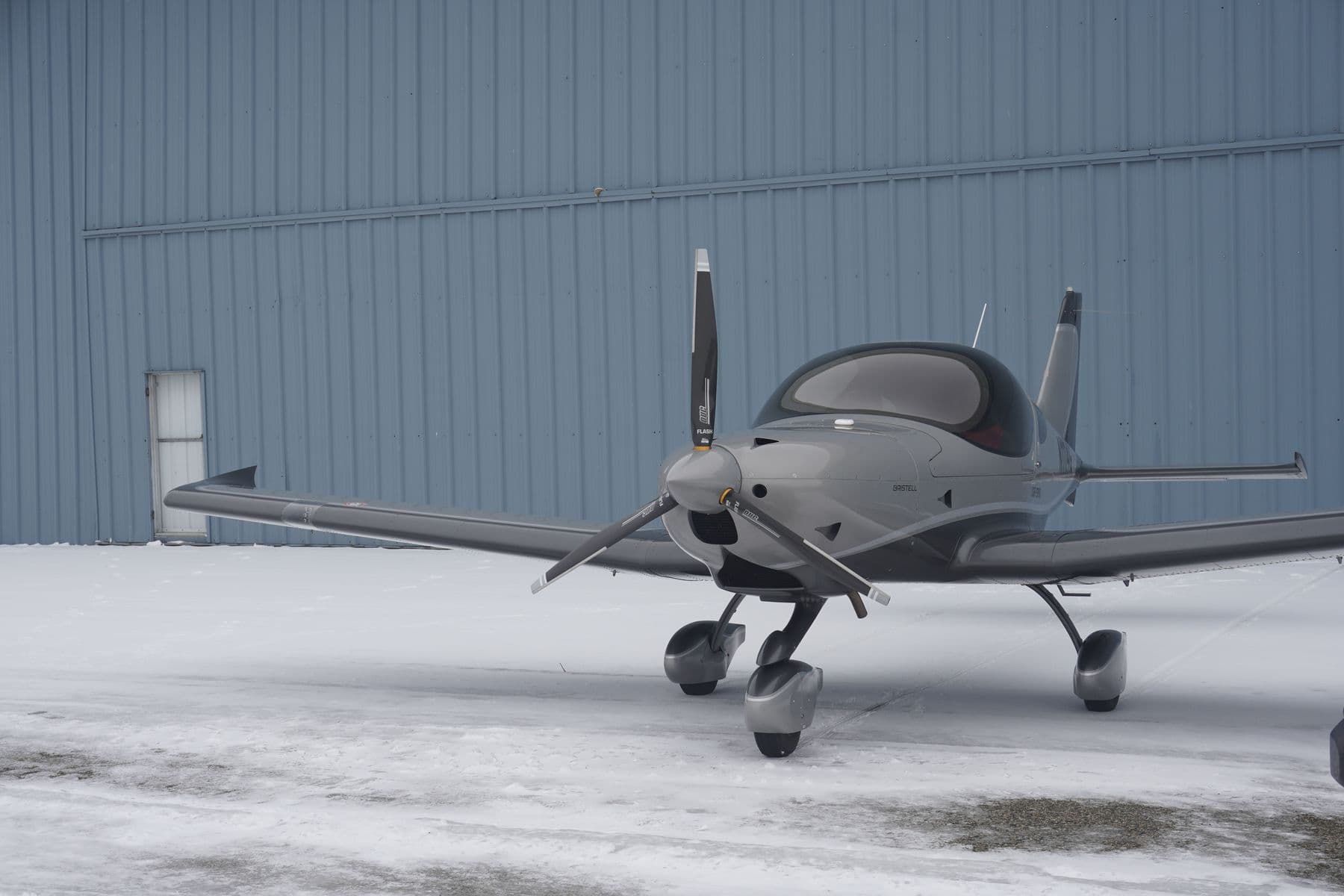 A silver Bristell in profile beside the blue hangar