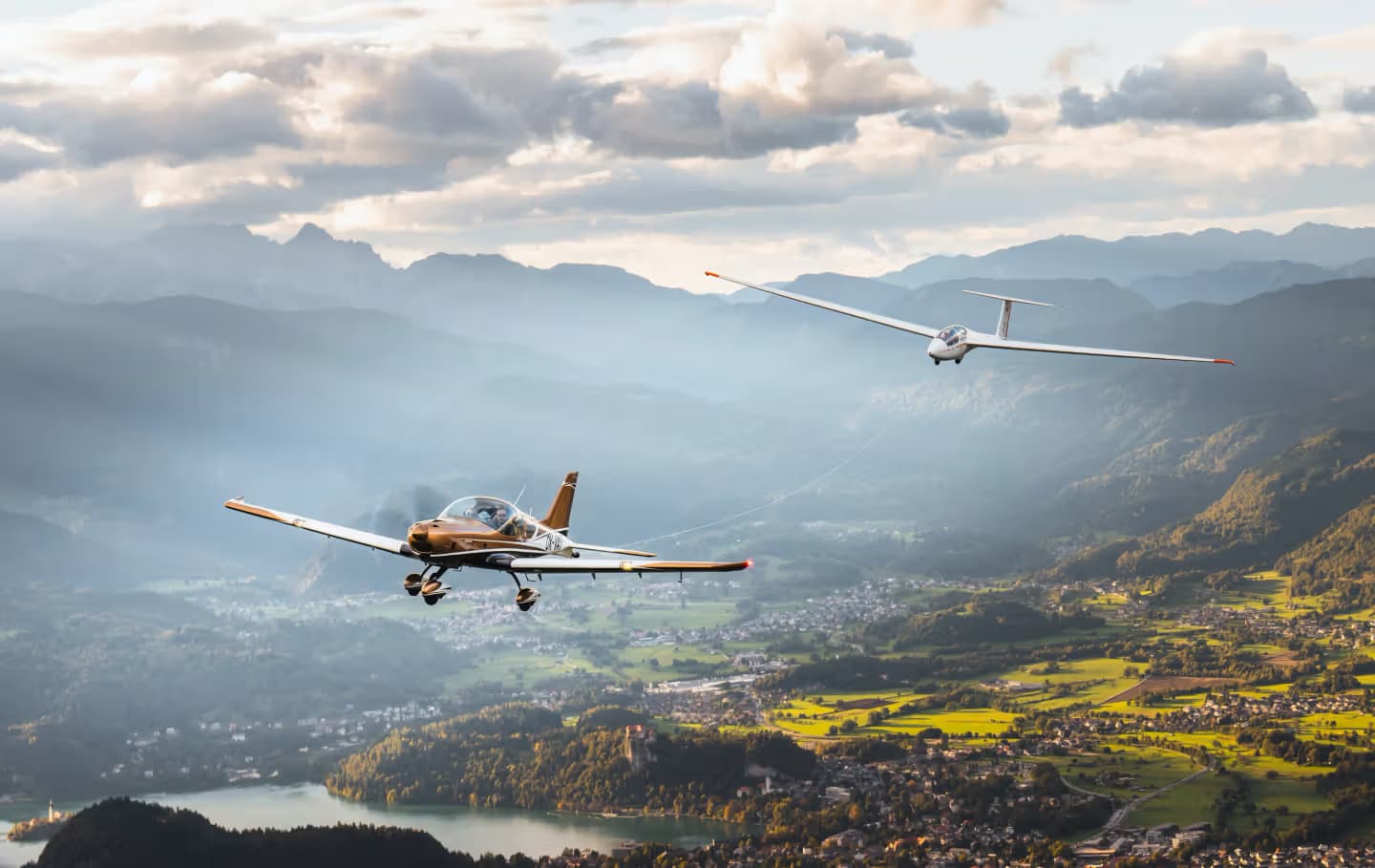 A Bristell towing a glider above an alpine valley in evening light