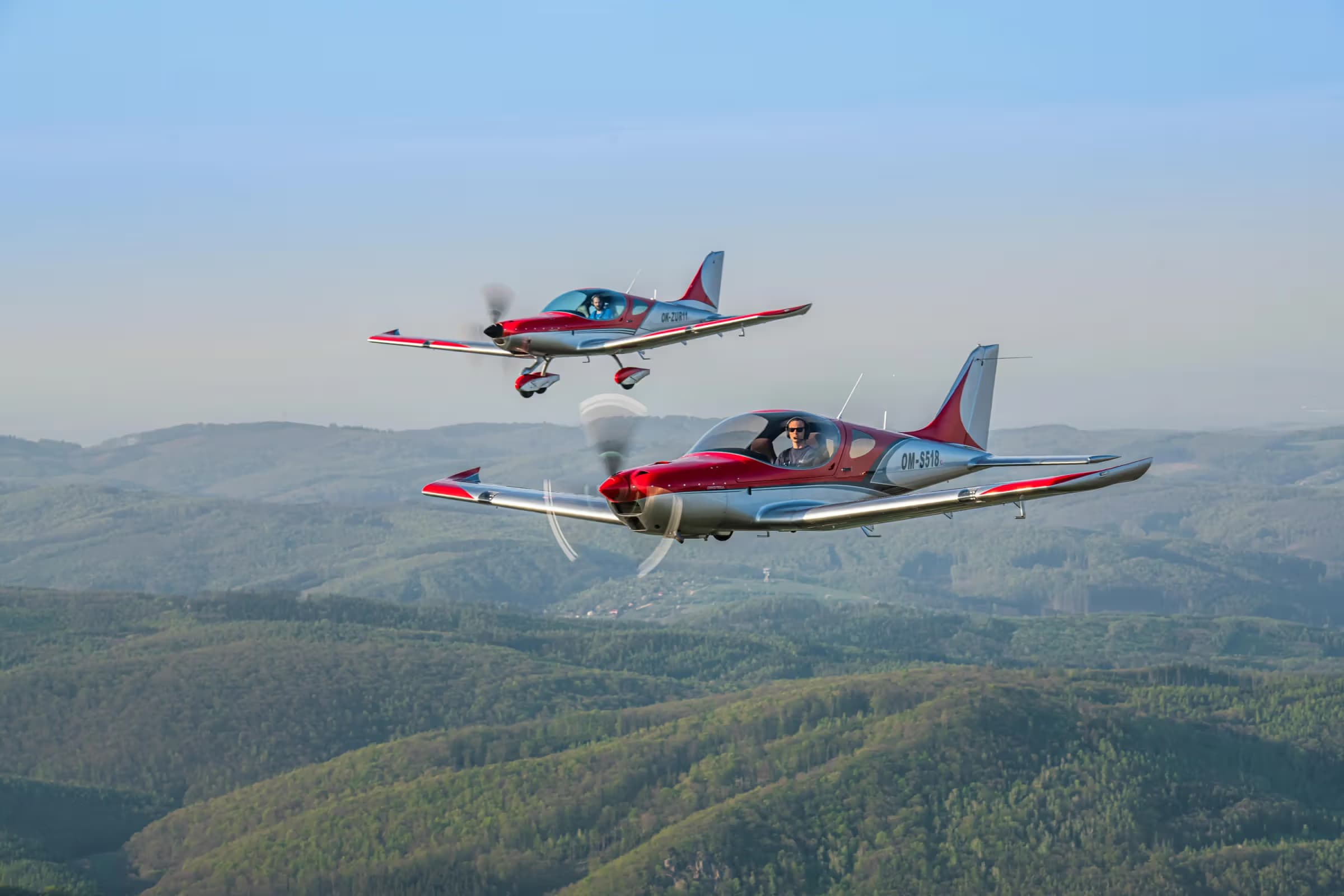 Two red and silver Bristells flying in formation above forested hills